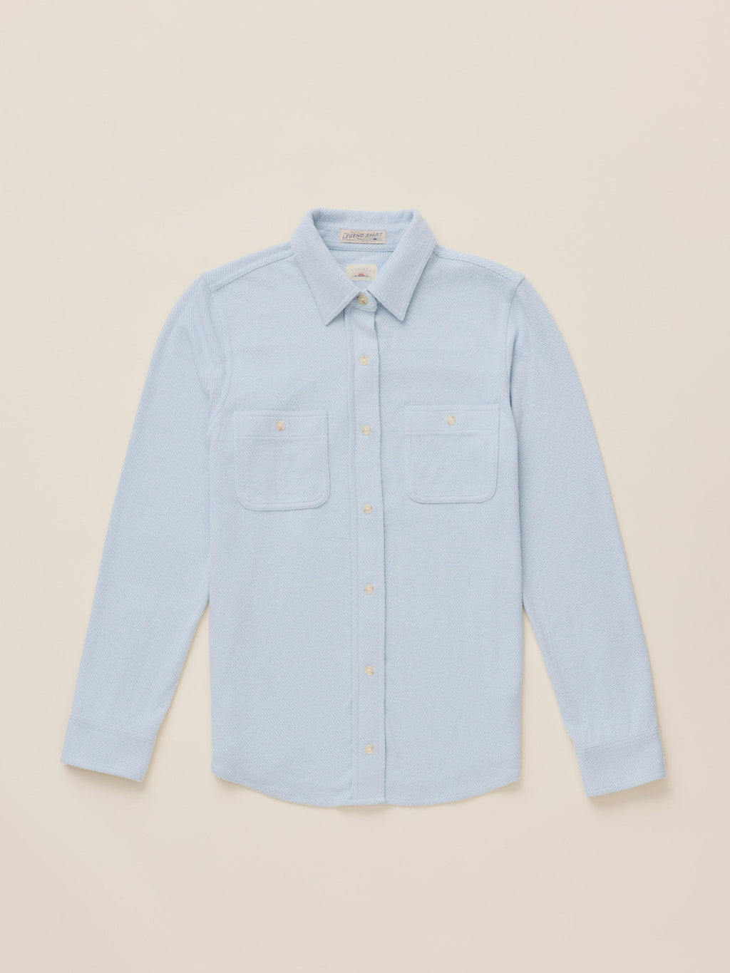 A light blue long-sleeve button-up shirt with two chest pockets, laid flat on a warm beige background.