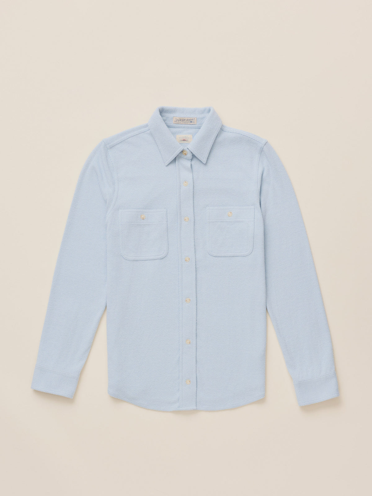 A light blue long-sleeve button-up shirt with two chest pockets, laid flat on a warm beige background.