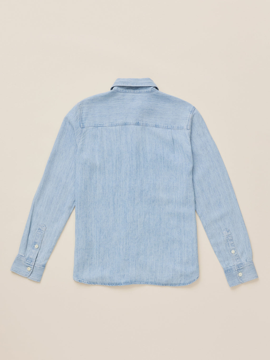 A light blue chambray long-sleeve button-up shirt shown from the back, laid flat on a warm beige background.