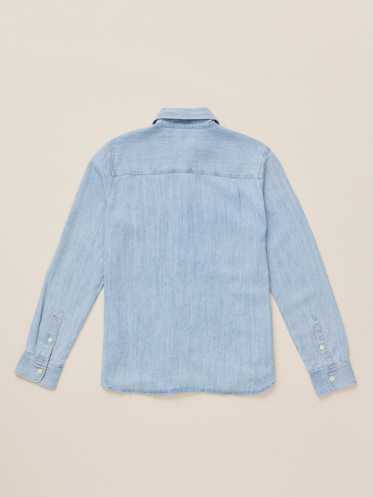 A light blue chambray long-sleeve button-up shirt shown from the back, laid flat on a warm beige background.